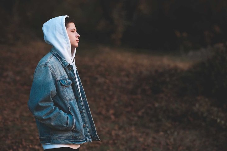 Handsome teen boy  wearing denim jacket and white hoodie standing outdoors over nature autumn background. Teenagerhood.