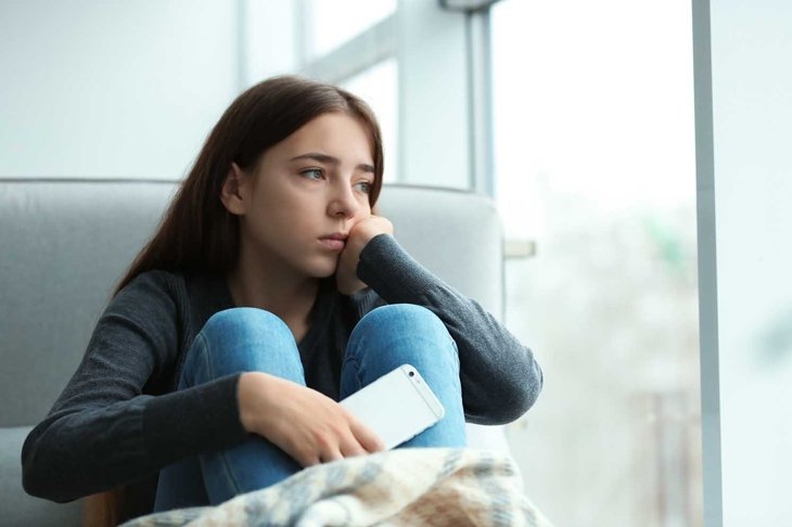 Upset teenage girl with smartphone sitting at window indoors. Space for text