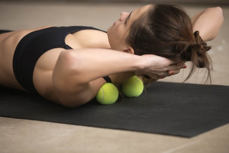 Lifestyle shot of young attractive female practicing self-massage technique applying tennis balls for neck and shoulder pain relief, working out on fitness mat on grey studio floor background, closeup