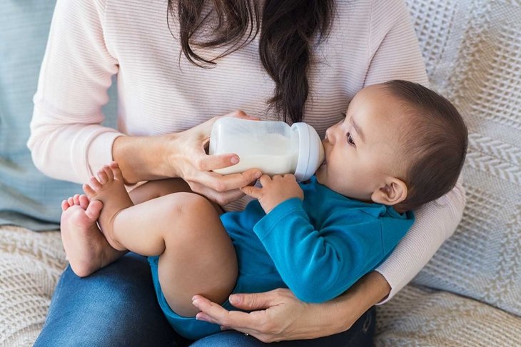 Little infant baby lying on mothers hand drinking milk from bottle. Hispanic loving mother feed her cute toddler while sitting on sofa.